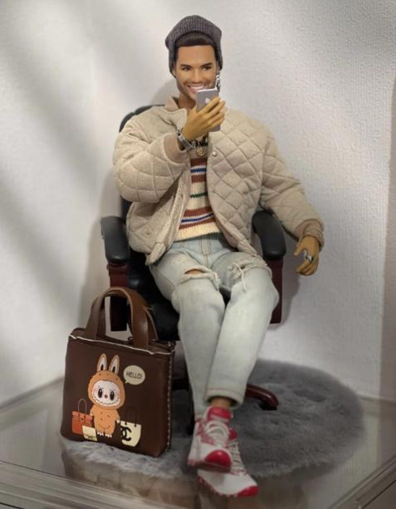 May include: A male fashion doll wearing a grey beanie, a beige quilted bomber jacket, a striped sweater, and light blue distressed jeans. The doll holds a small silver smartphone and sits on a black chair next to a brown tote bag with a cartoon graphic.