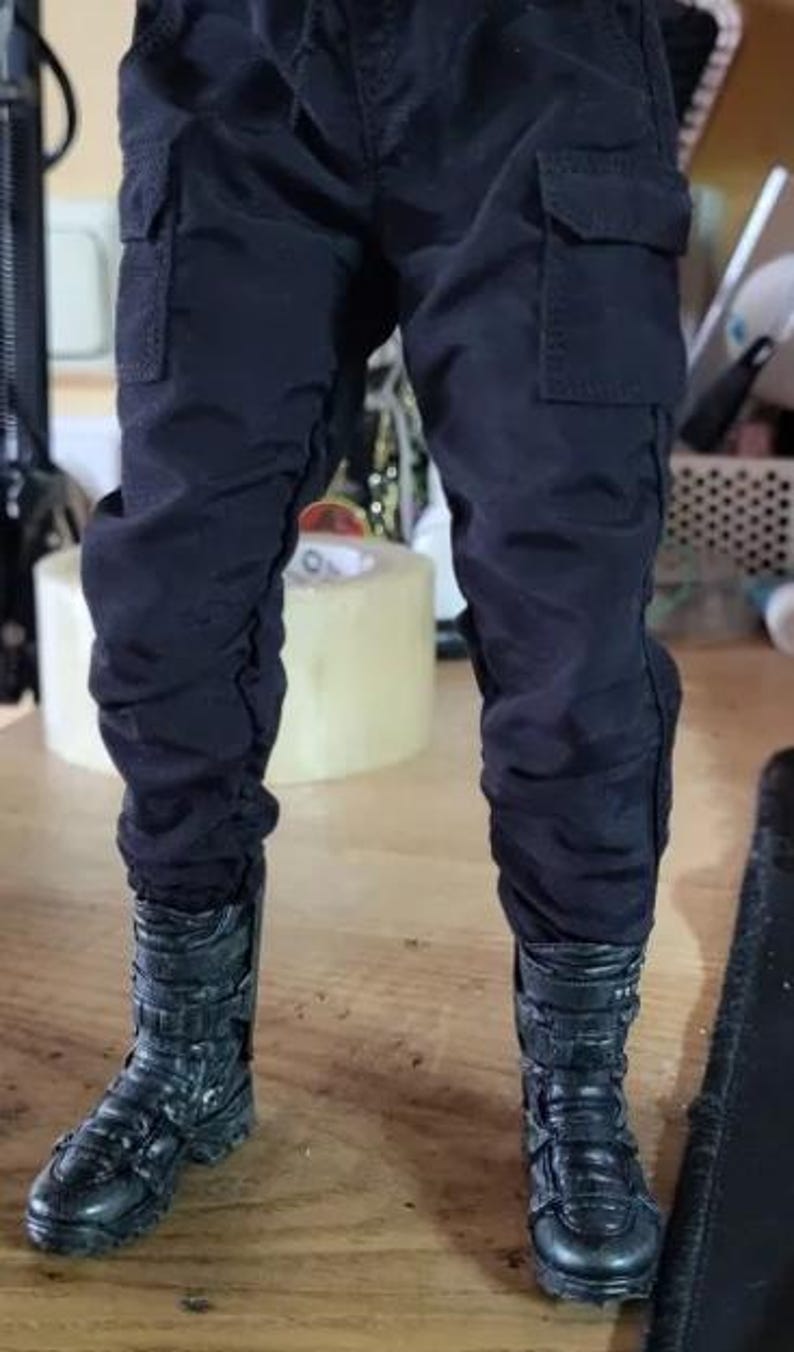 May include: Black cargo pants with multiple pockets and black combat boots. The pants have a slightly wrinkled appearance. The boots have a textured design and multiple straps.