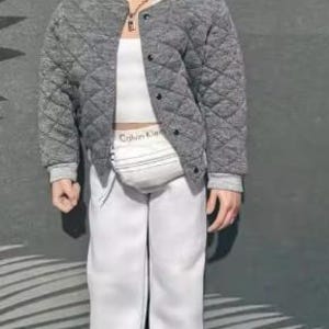 May include: A doll with brown hair wearing a grey quilted bomber jacket over a white crop top and white sweatpants. The doll features a white fanny pack with the text Calvin Klein, a silver chain necklace, and white trainers with red accents.