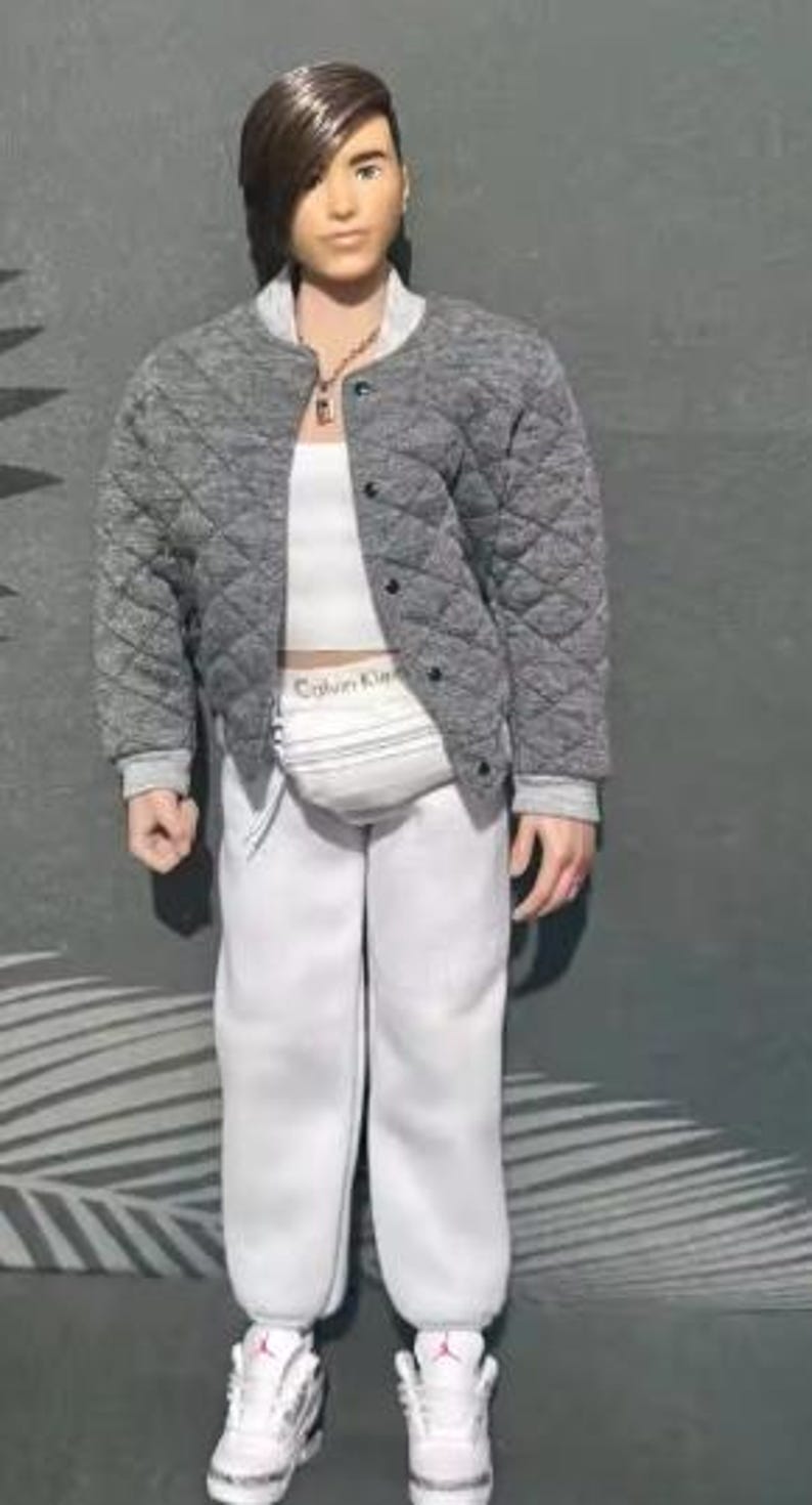 May include: A doll with brown hair wearing a grey quilted bomber jacket over a white crop top and white sweatpants. The doll features a white fanny pack with the text Calvin Klein, a silver chain necklace, and white trainers with red accents.