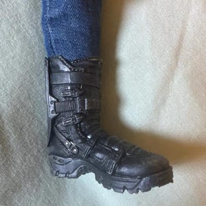 May include: Black combat boot with multiple buckles and straps. The boot has a rugged sole and is worn with blue denim jeans. The boot appears to be made of a durable material.