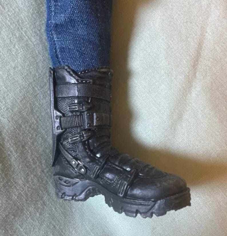 May include: Black combat boot with multiple buckles and straps. The boot has a rugged sole and is worn with blue denim jeans. The boot appears to be made of a durable material.