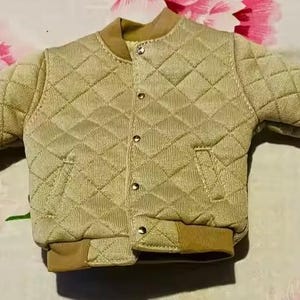 May include: A beige quilted bomber jacket featuring a diamond-stitched pattern, a ribbed collar, ribbed cuffs, and a front closure with metallic snap buttons. The jacket includes two side pockets and a solid tan hem at the bottom of the garment.