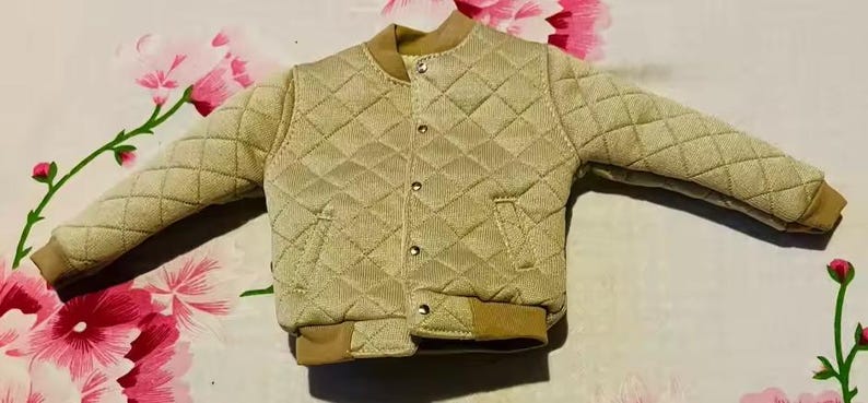 May include: A beige quilted bomber jacket featuring a diamond-stitched pattern, a ribbed collar, ribbed cuffs, and a front closure with metallic snap buttons. The jacket includes two side pockets and a solid tan hem at the bottom of the garment.