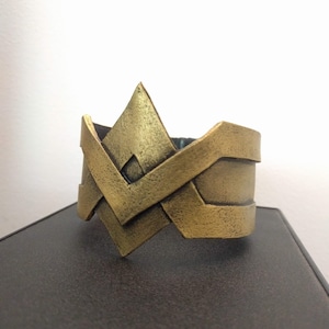 May include: A gold-colored metal cuff bracelet with a geometric design. The design features a large, pointed diamond shape with smaller, angled lines extending from the sides.