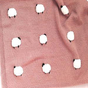 Może przedstawiać: Miękki, różowy, dziergany kocyk dla niemowląt z białymi aplikacjami owiec. Kocyk wykonany jest z miękkiego, przytulnego materiału i idealnie nadaje się do przytulania.