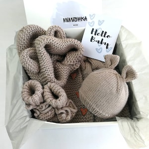 Könnte beinhalten: Ein beiges Strick-Babygeschenkset in einer weißen Box. Das Set enthält eine Decke, Babyschuhe, einen Pullover mit herzförmigen Knöpfen und ein Stofftier mit ohrenartigen Vorsprüngen. Eine Karte mit dem Text "Hello Baby" ist ebenfalls enthalten.