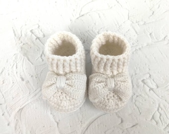 Pode incluir: Um par de sapatinhos de bebé de crochê branco com um laço na frente de cada sapatinho.