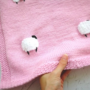 Könnte beinhalten: Eine rosa gestrickte Babydecke mit weißen, flauschigen Schaf-Applikationen. Die Decke hat einen rosa gestrickten Rand.
