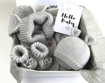 May include: A gray knitted baby gift set in a white box. The set includes a blanket, booties, a sweater, and a hat with bunny ears. A card with the words "Hello Baby" is also included.