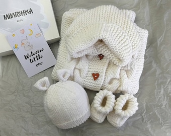 May include: A white knitted baby gift set including a cardigan with heart-shaped buttons, a hat with ear details, and booties. A card with the text "Welcome little one" and a box with the brand name "Mimishka Kids" are also included.