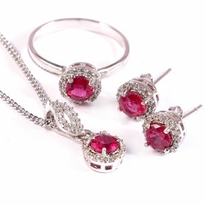 May include: A set of ruby and diamond jewelry featuring a ring, a pendant necklace, and a pair of earrings. The jewelry is made of white gold and features a red ruby gemstone surrounded by smaller diamonds.