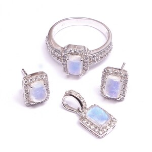 Puede incluir: Un conjunto de joyer&iacute;a compuesto por un anillo, un par de pendientes de bot&oacute;n y un colgante de plata. Cada pieza presenta un cabuj&oacute;n de piedra lunar rectangular e iridiscente rodeado por un halo de peque&ntilde;as piedras blancas brillantes.