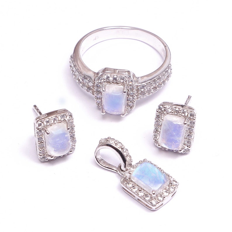 Puede incluir: Un conjunto de joyer&iacute;a compuesto por un anillo, un par de pendientes de bot&oacute;n y un colgante de plata. Cada pieza presenta un cabuj&oacute;n de piedra lunar rectangular e iridiscente rodeado por un halo de peque&ntilde;as piedras blancas brillantes.