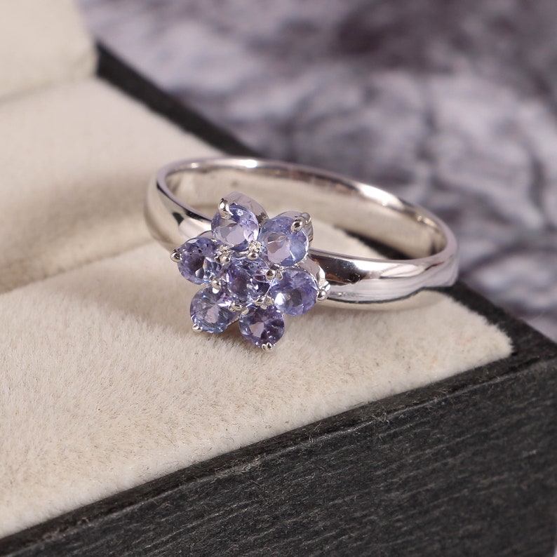 Puede incluir: Un anillo de plata con un dise&ntilde;o floral con piedras preciosas de color p&uacute;rpura claro. El anillo se presenta en una caja de joyer&iacute;a negra con un interior beige claro, sobre un fondo borroso.