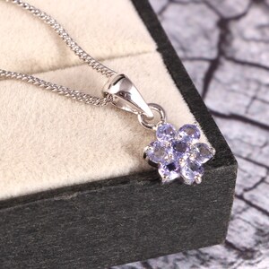 Puede incluir: Un collar de plata con un colgante de flor. El colgante est&aacute; formado por peque&ntilde;as piedras preciosas de color p&uacute;rpura claro dispuestas en un patr&oacute;n floral. El collar se muestra en una caja de joyer&iacute;a negra con un interior beige claro.