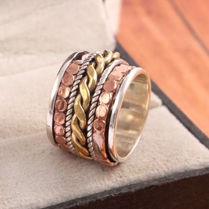 Peut inclure: Une bague large composée d'un fil torsadé doré central, entouré de deux cordes tressées argentées et de deux bandes extérieures à la texture cuivrée martelée. La bague présente un intérieur argenté poli et des bords lisses argentés.