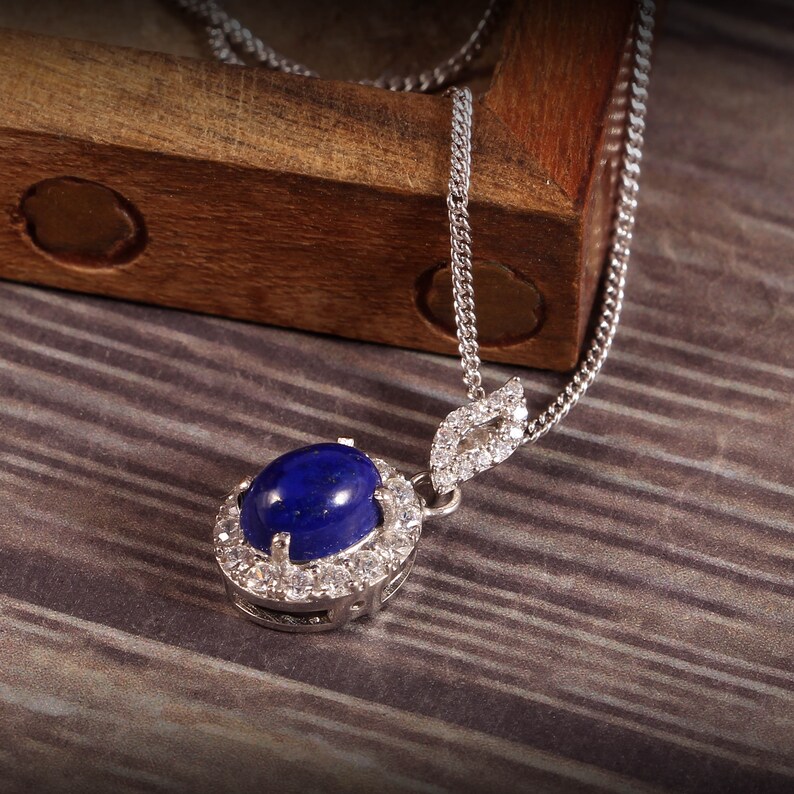 Puede incluir: Un collar de cadena de plata con un colgante que presenta una gran piedra ovalada de lapisl&aacute;zuli rodeada por un halo de piedras de circonita c&uacute;bica transparentes m&aacute;s peque&ntilde;as.