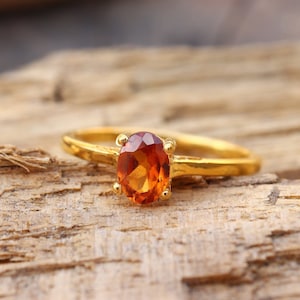 May include: A gold ring with a large, oval-shaped, orange gemstone. The ring is sitting on a piece of wood.