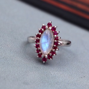 May include: A silver ring with a large, oval-shaped moonstone set in the center. The moonstone is surrounded by a halo of small, red rubies.