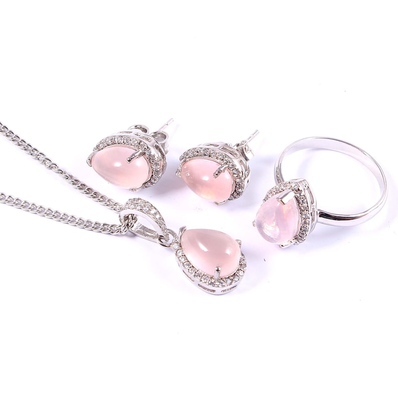 May include: A jewelry set featuring a necklace, earrings, and a ring. Each piece has a pear-shaped, light pink gemstone surrounded by a silver-colored metal setting with small, clear accent stones. The necklace has a delicate chain.