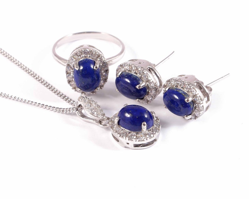 Puede incluir: Un conjunto de joyas de plata con un collar, un anillo y unos pendientes. Cada pieza presenta una piedra ovalada de lapisl&aacute;zuli azul rodeada por un halo de piedras m&aacute;s peque&ntilde;as y transparentes.