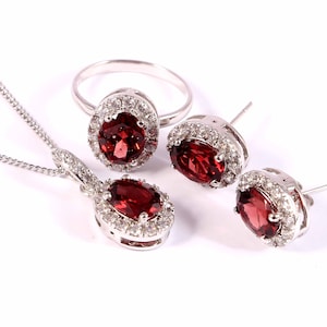 May include: A silver jewellery set with a red gemstone pendant, ring, and earrings. Each piece features a red oval gemstone surrounded by a halo of smaller white gemstones.