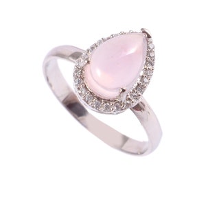 May include: A silver ring featuring a pear-shaped, light pink gemstone surrounded by a halo of small, clear stones. The ring's band is a simple, polished silver. The gemstone is the focal point of the ring.