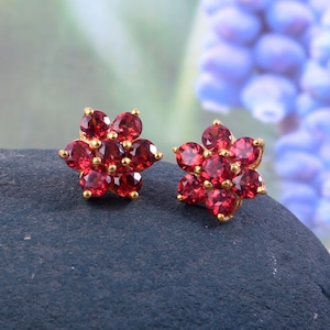 May include: A pair of gold flower-shaped earrings with red gemstones. Each earring has nine red gemstones set in a gold flower design.