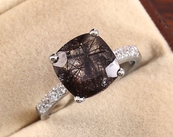 May include: A silver ring with a large, square-shaped, dark gray gemstone with black inclusions. The ring has a band of small, clear gemstones.