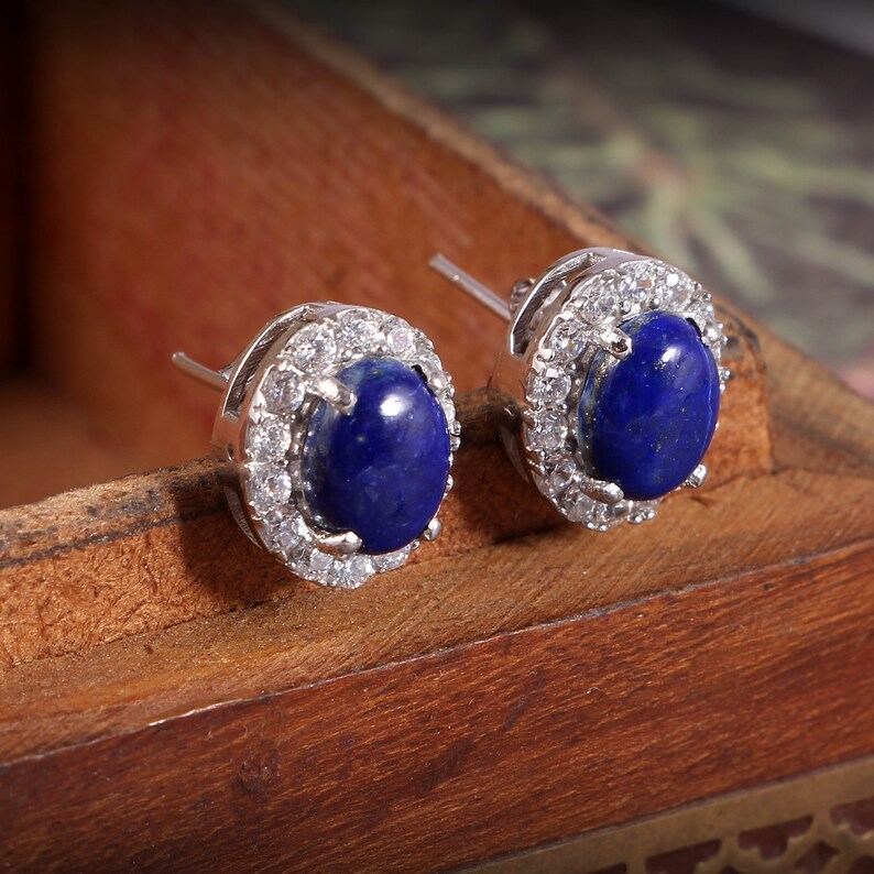 Puede incluir: Un par de pendientes de plata con piedras ovaladas de lapisl&aacute;zuli azul rodeadas por un halo de piedras de circonita c&uacute;bica transparentes.