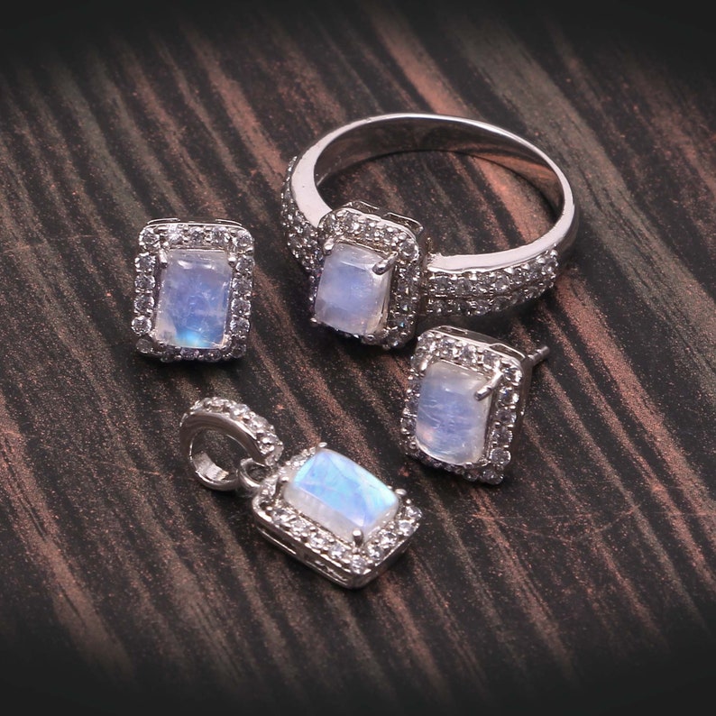 Puede incluir: Un conjunto de joyer&iacute;a compuesto por un anillo, un par de pendientes y un colgante de tono plateado. Cada pieza presenta una piedra lunar rectangular iridiscente rodeada por un halo de peque&ntilde;as piedras de circonita c&uacute;bica blancas.