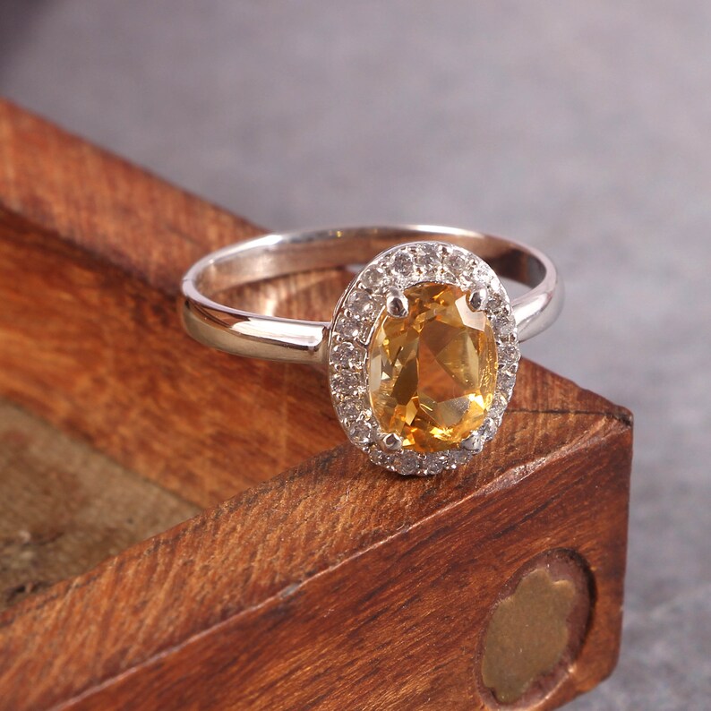May include: A silver ring with a large oval-shaped topaz gemstone surrounded by smaller clear gemstones. The ring is sitting on a wooden surface.