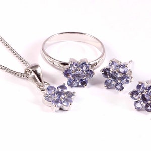 Puede incluir: Un conjunto de joyas que incluye un collar con colgante, un anillo y pendientes. Cada pieza tiene un engaste plateado y est&aacute; adornada con un grupo de peque&ntilde;as piedras preciosas de color p&uacute;rpura claro dispuestas en forma de flor. El collar tiene una cadena fina.