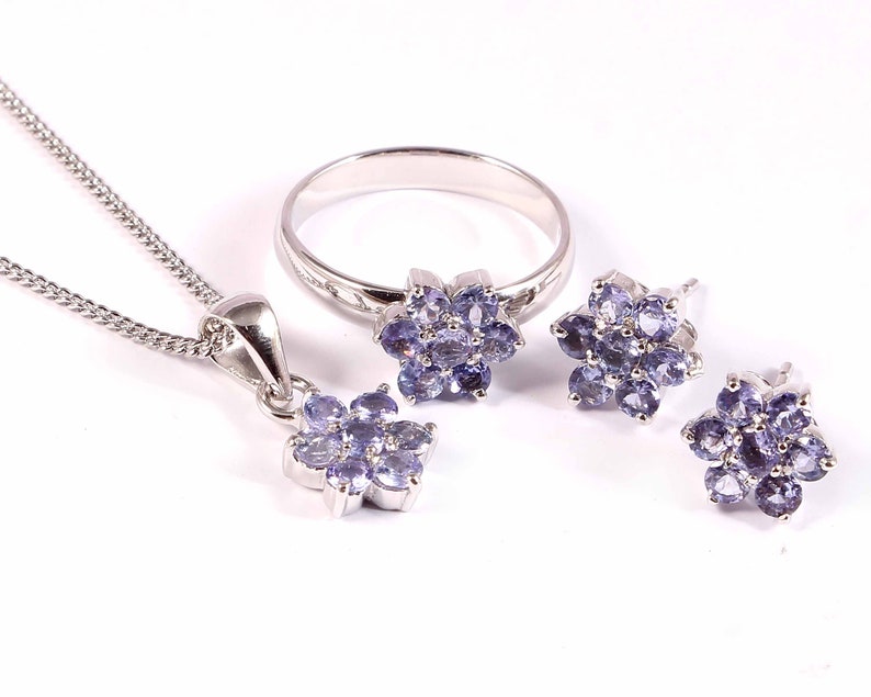 Puede incluir: Un conjunto de joyas que incluye un collar con colgante, un anillo y pendientes. Cada pieza tiene un engaste plateado y est&aacute; adornada con un grupo de peque&ntilde;as piedras preciosas de color p&uacute;rpura claro dispuestas en forma de flor. El collar tiene una cadena fina.