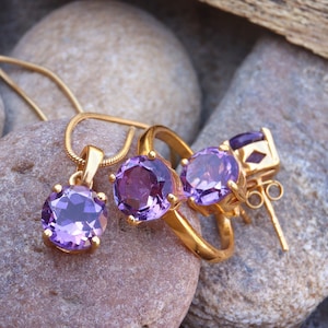 May include: A set of gold jewellery featuring three amethyst gemstones. The set includes a ring, a pendant, and a pair of earrings.