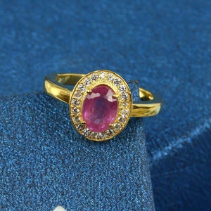 Puede incluir: Un anillo de oro con un rubí ovalado rodeado de pequeñas piedras preciosas transparentes. La banda del anillo es dorada. El anillo se muestra sobre un fondo azul. El rubí es el punto focal del anillo.