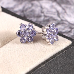 Puede incluir: Un par de pendientes de plata, cada uno con un dise&ntilde;o floral. Cada flor est&aacute; compuesta por varias piedras preciosas ovaladas de color p&uacute;rpura claro. Los pendientes se exhiben en una caja de joyer&iacute;a negra con interior beige.