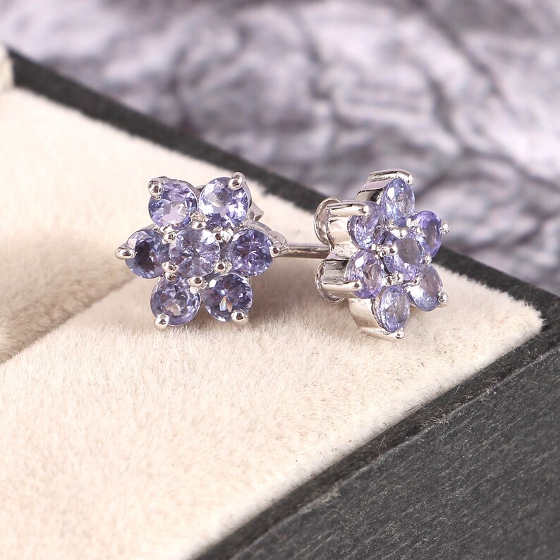 Puede incluir: Un par de pendientes de plata, cada uno con un dise&ntilde;o floral. Cada flor est&aacute; compuesta por varias piedras preciosas ovaladas de color p&uacute;rpura claro. Los pendientes se exhiben en una caja de joyer&iacute;a negra con interior beige.