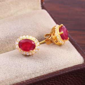 以下が含まれることがあります： 楕円形の赤いルビーの宝石を、より小さな白い宝石のハローで囲んだ、金色のイヤリングのペアです。