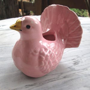 Könnte beinhalten: Eine rosafarbene Keramik-Vogelfigur mit gelbem Schnabel und einem gekräuselten Schwanz. Der Vogel ist als Pflanzgefäß konzipiert.