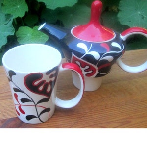 Könnte beinhalten: Eine weiße Keramiktasse mit einem schwarzen und roten Blumenmuster und eine passende Teekanne mit einem roten Deckel und einem schwarzen und weißen Blumenmuster.