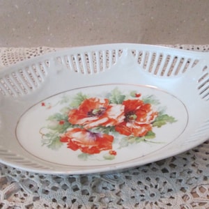 Könnte beinhalten: Weiße Porzellanservierschale mit durchbrochenen Seiten und Griffen. Die Schale zeigt ein florales Design mit roten Mohnblumen und grünen Blättern.