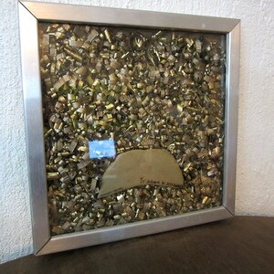 Könnte beinhalten: Ein gerahmtes Kunstwerk mit einem silbernen Metallrahmen und einer Glasscheibe, die mit goldenen und silbernen Metallic-Konfetti gefüllt ist. Das Konfetti ist in einem zufälligen Muster angeordnet und scheint auf das Glas geklebt zu sein. Das Kunstwerk ist unten rechts mit "Sr. Adorno" signiert.