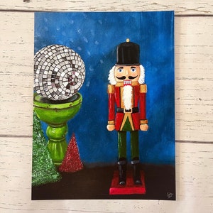 May include: A painting of a Christmas scene featuring a nutcracker soldier with a black hat and red coat. A disco ball sits on a green pedestal, and two small Christmas trees are also in the painting. The background is a dark blue color.