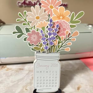 May include: A papercraft calendar for January 2026, shaped like a mason jar filled with colorful paper flowers. The flowers include pink, peach, and lavender blooms with green leaves. The calendar is on a black stand.
