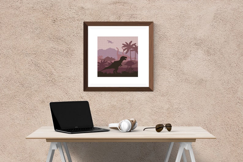 May include: A framed print of a dinosaur silhouette in a pink and purple landscape with palm trees. The print is hanging on a beige wall above a wooden desk with a laptop, headphones, and sunglasses.