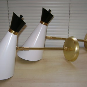 Pode incluir: Dois candeeiros de parede cónicos brancos e pretos com braços e bases de metal dourado. Os candeeiros são ajustáveis e podem ser inclinados para cima ou para baixo.