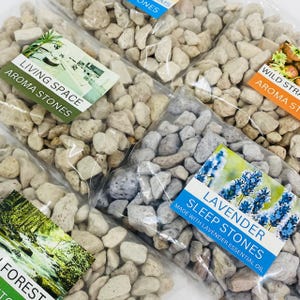May include: Multiple clear bags filled with small stones in various shades of grey and beige. Each bag features a label with text including "Living Space Aroma Stones" and "Sleep Stones Made with Lavender Essential Oil".