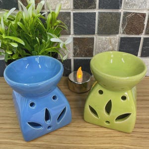 May include: Two ceramic oil warmers, one blue and one green, are displayed. Each warmer has a bowl and a base with leaf-shaped cutouts. A small, lit candle sits between them. A green plant is in the background.